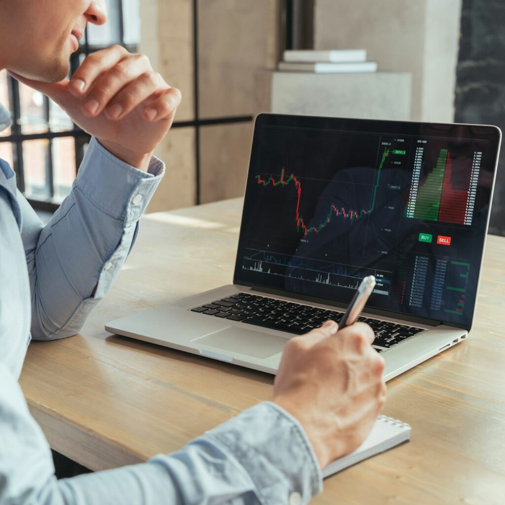 Cropped image of crypto analyst in front of laptop with chart, writing trading strategy in notepad